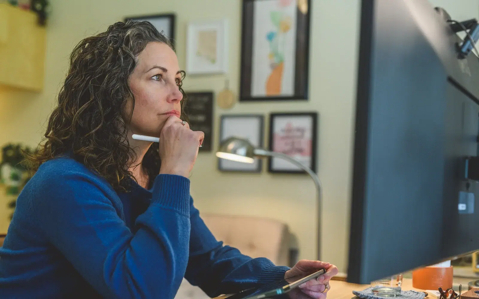 Heather Young in a blue sweater sits at her desk with a stylus in hand, looking thoughtfully toward a monitor, framed prints and a desk lamp on the wall behind her.