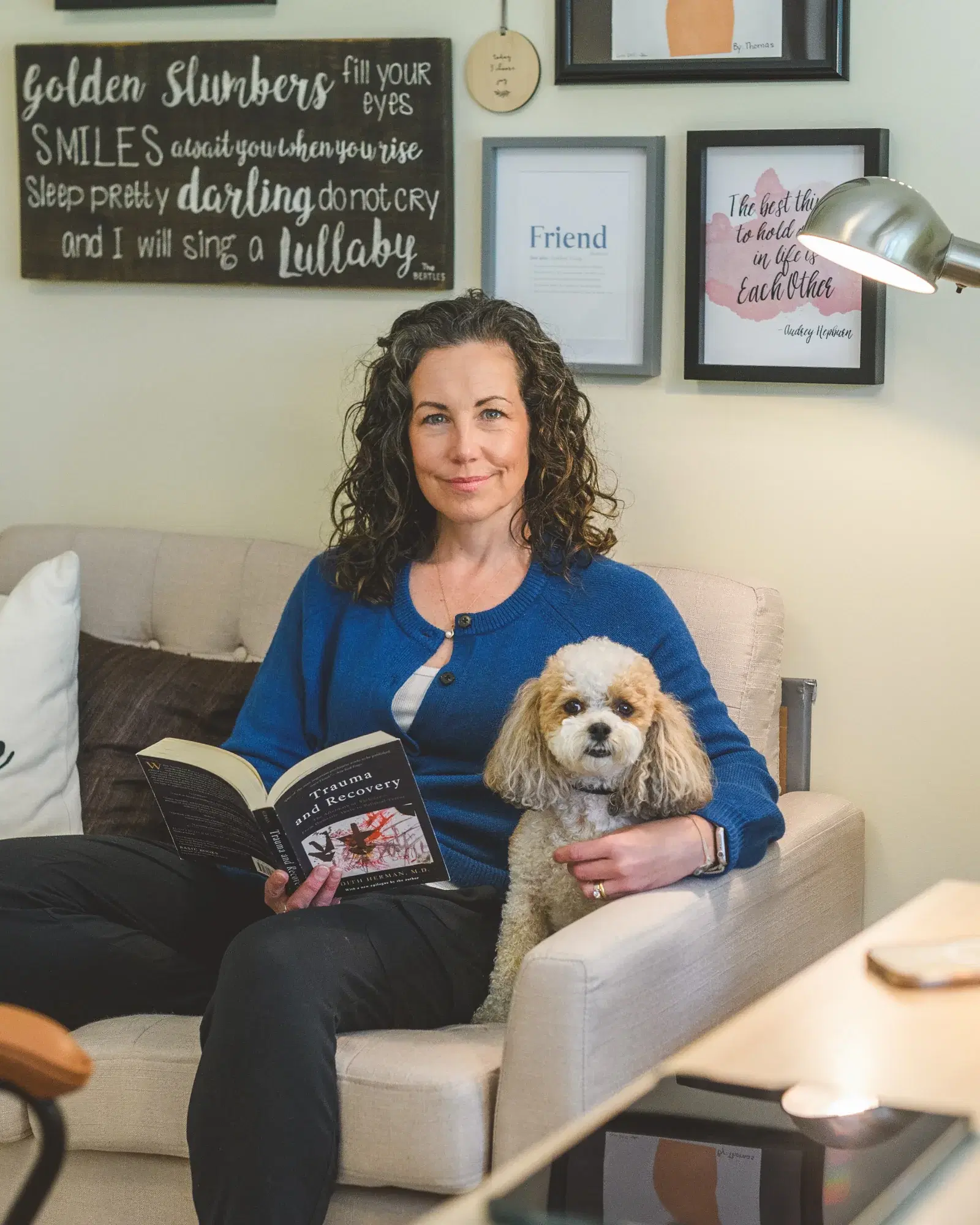 Heather Young, MSW RSW PMH-C, in a soft blue blouse, seated with her arms resting on a table, looking warmly toward the camera, with framed art on the wall behind her.