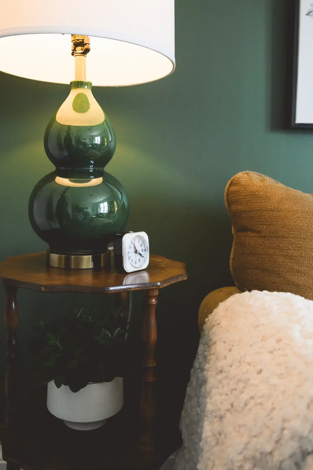 A close detail of the office: a green ceramic table lamp on a wooden side table next to a mustard armchair with a small clock and a sheepskin throw.