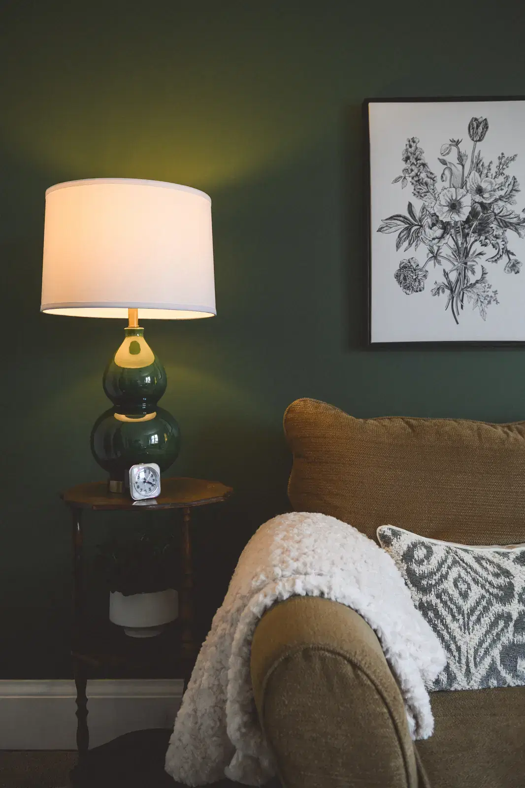A soft detail: a green ceramic table lamp glowing against a deep green wall, beside a small clock, a knit throw, and a black and white botanical print.