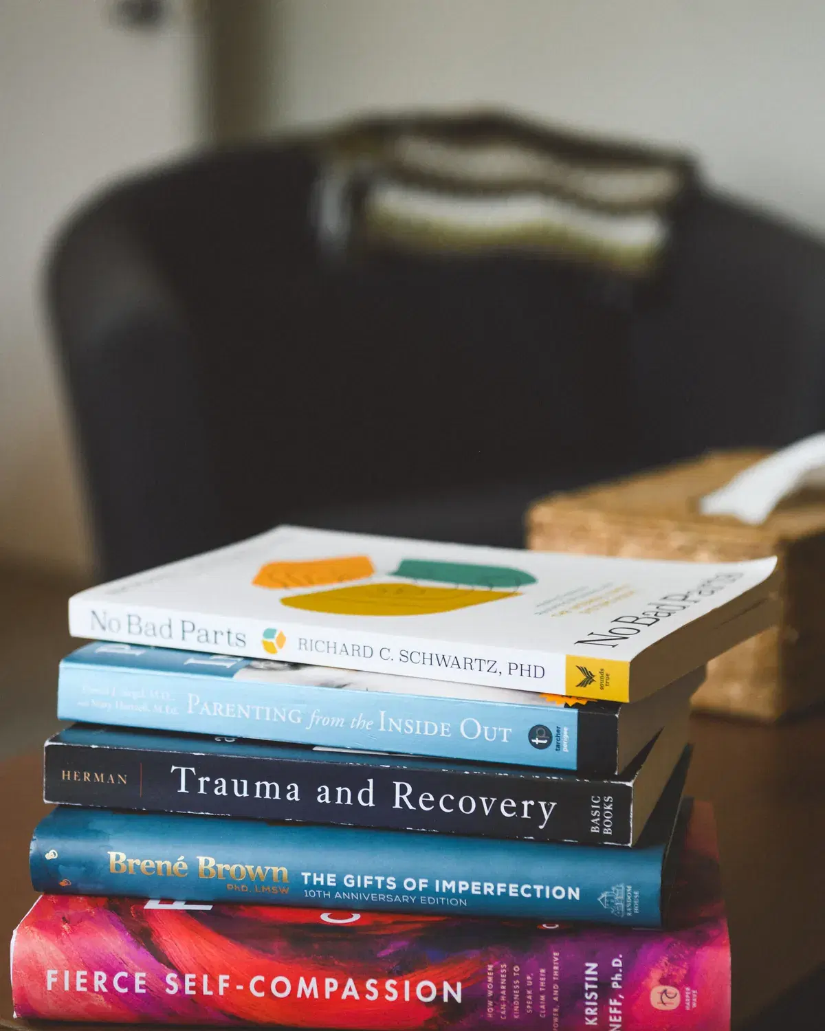A stack of well-used books on a low table in a counselling room, spines including No Bad Parts, Parenting from the Inside Out, and Fierce Self-Compassion.