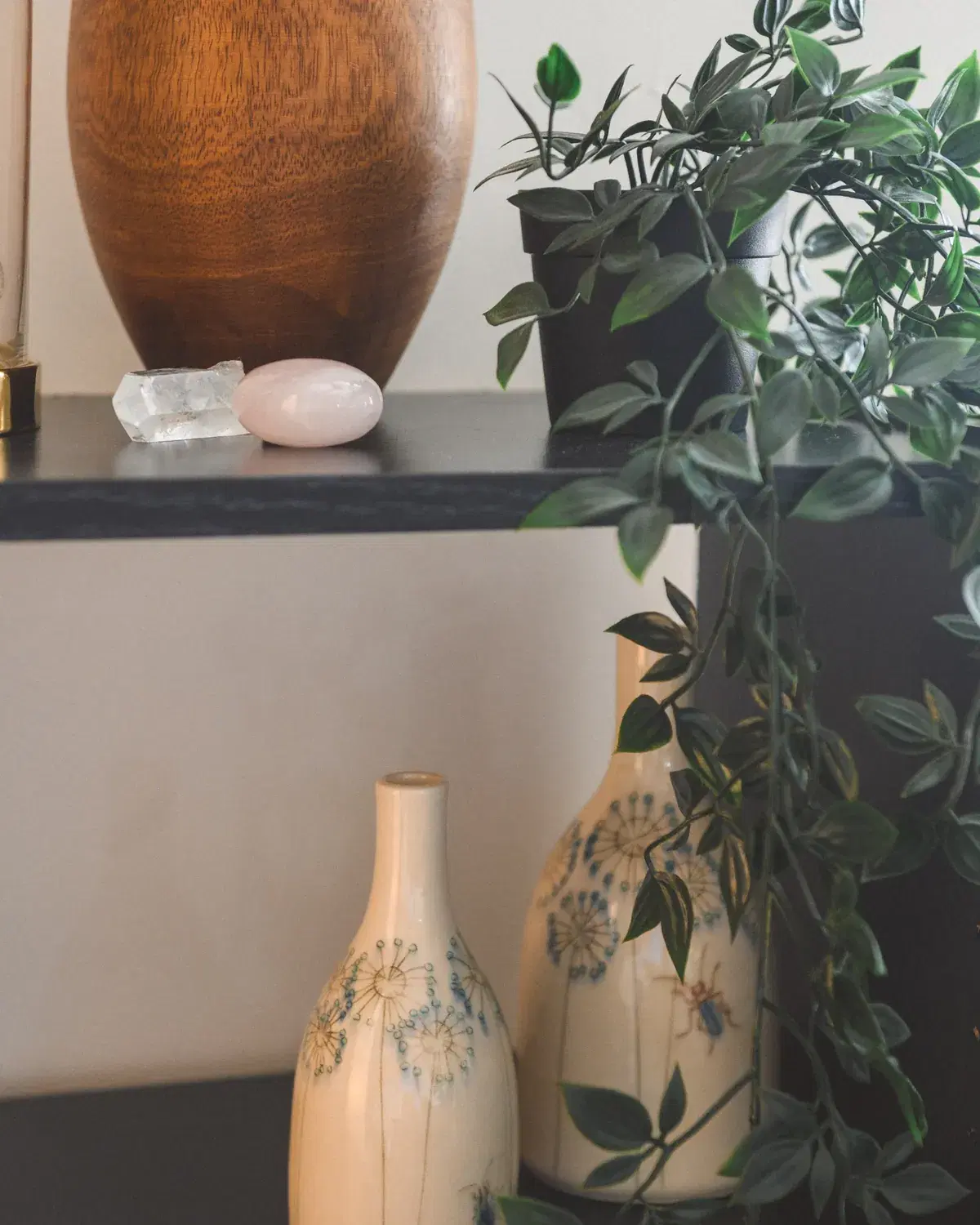 A quiet shelf detail with trailing green plants, a smooth wooden vessel, and softly patterned ceramics in gentle daylight.